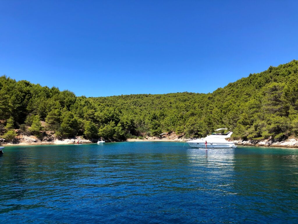 Clear blue bay with pine-covered hills, a rocky shoreline, and a white yacht anchored near the shore; a person stands on the deck.