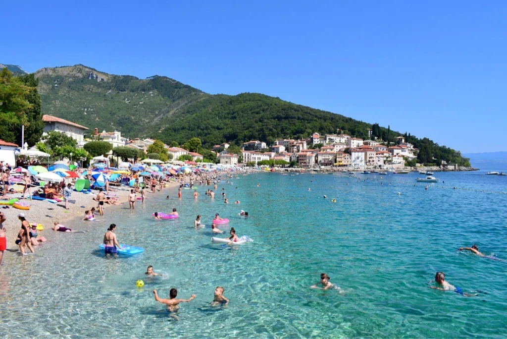 Crowded pebble beach with colorful umbrellas and sunbathers, turquoise water, and green hills rising behind a seaside town.