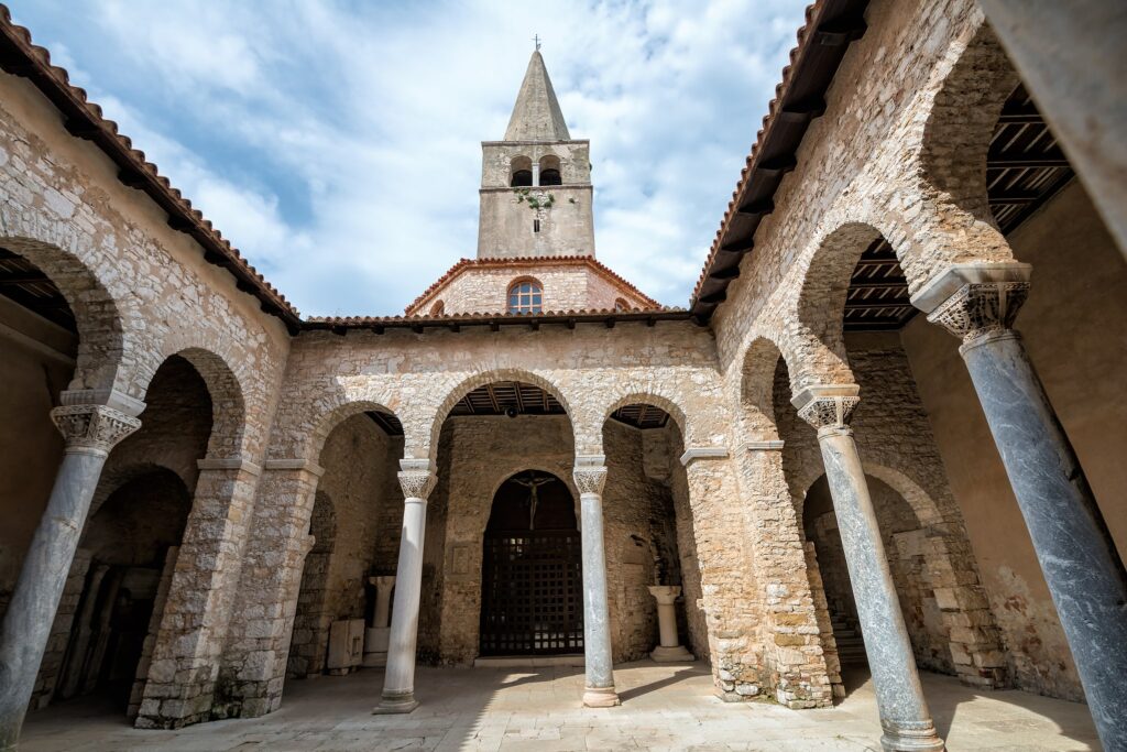 Interior of a historic stone cloister with arches and slender columns, a church tower rising above under a blue sky.